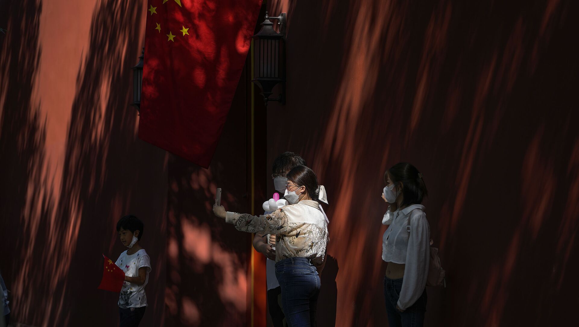 A couple wearing face masks to help protect from the coronavirus pose for a selfie near a national flag on a wall cast with shadows from trees on National Day in Beijing, Friday, Oct. 1, 2021. Hundreds of thousands domestic tourists flock to the square to celebrate the 72nd National Day of the People's Republic of China over a week-long holiday. (AP Photo/Andy Wong) - 俄罗斯卫星通讯社, 1920, 02.10.2021