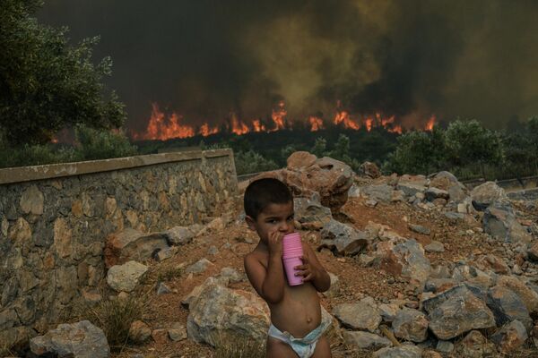 森林火災在希臘雅典周邊燃燒。圖為阿吉奧斯·哈拉拉姆波斯村一個孩子在自家院子里與身後燃燒的山火。 - 俄羅斯衛星通訊社