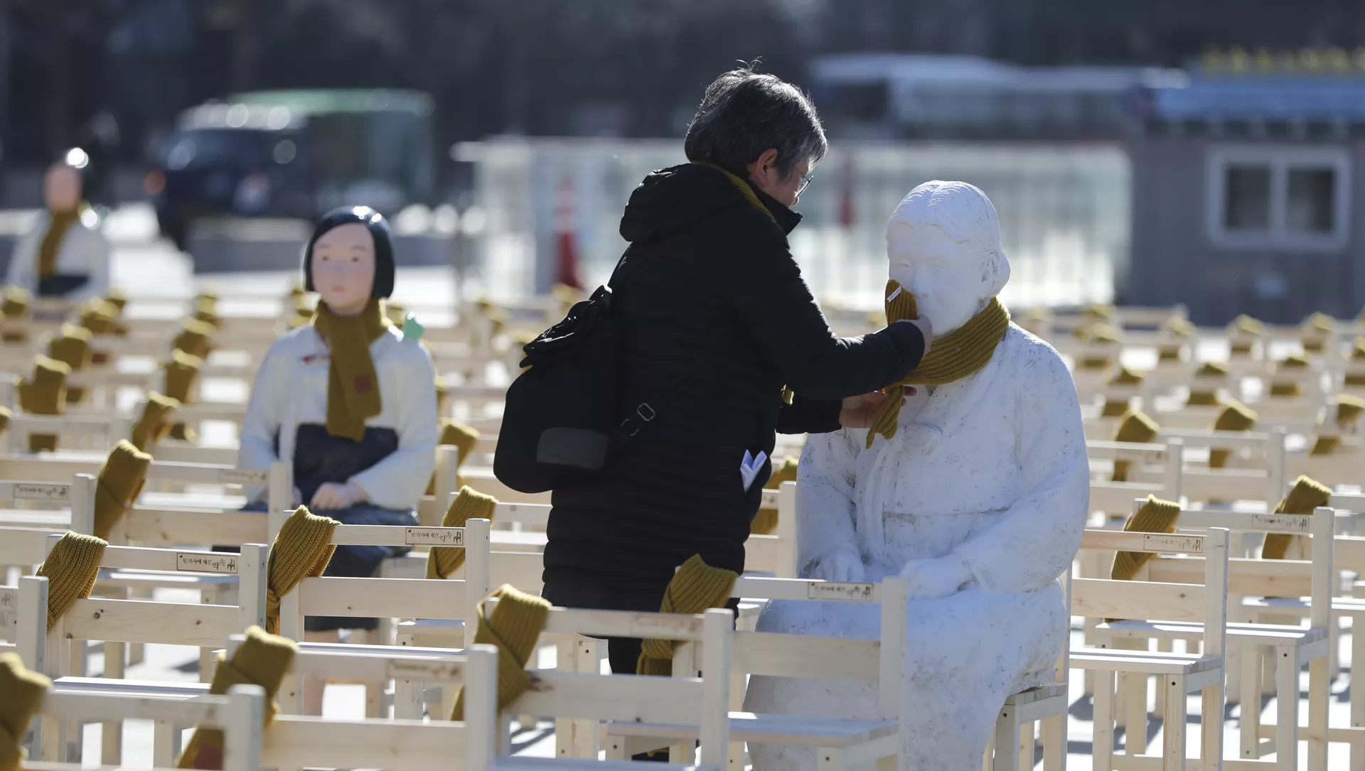 A woman puts a scarf on a statue of a comfort woman sitting in a installation of empty chairs symbolizing the victims in Seoul