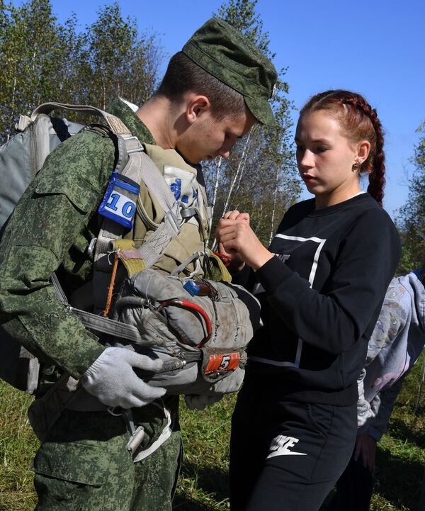 一名来自阿尔谢尼耶夫市航空俱乐部的女孩在滨海边疆区诺沃瑟索耶夫克村附近的野战机场跳伞前帮助乌苏里斯克苏沃洛夫军事学校的学员穿上降落伞 - 俄罗斯卫星通讯社