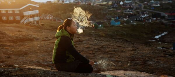 年轻的女孩坐在石头上，在落日的余晖中抽烟，塔西拉克镇（Tasiilaq Town），格陵兰岛 - 俄罗斯卫星通讯社